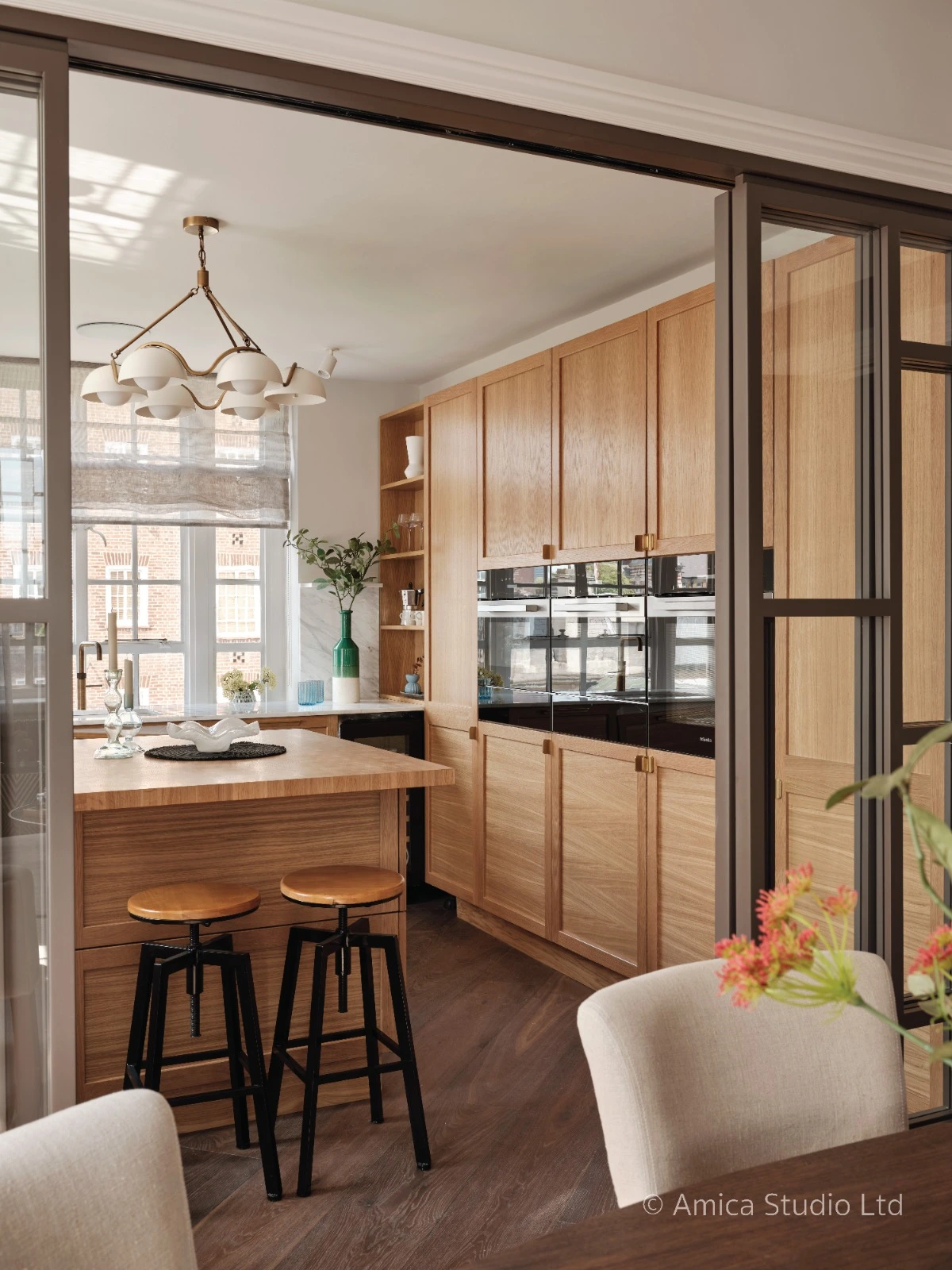 Bespoke oak kitchen viewed through bronze steel-framed glazed partition doors, with marble splashback and brass chandelier